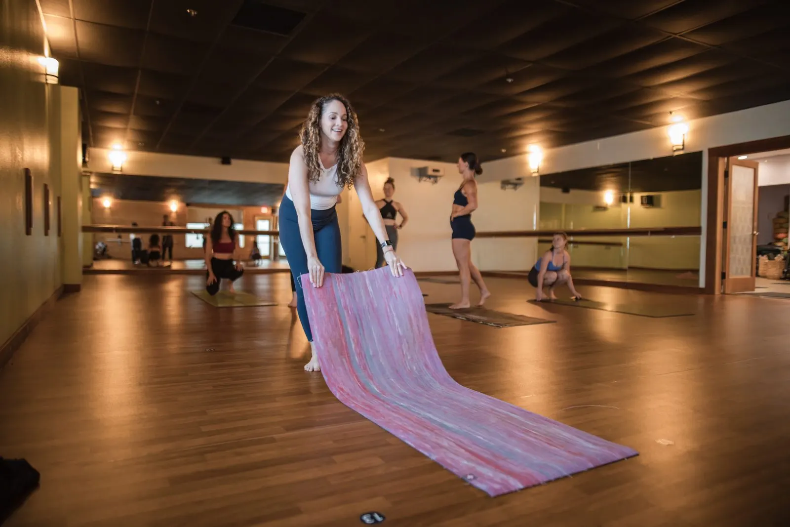 An instructor smiles as she rolls out her mat, while students set up behind her